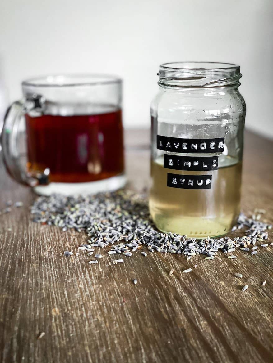 Lavender simple Syrup in jar. Coffee cup behind it all sitting on a bed of lavender buds. 