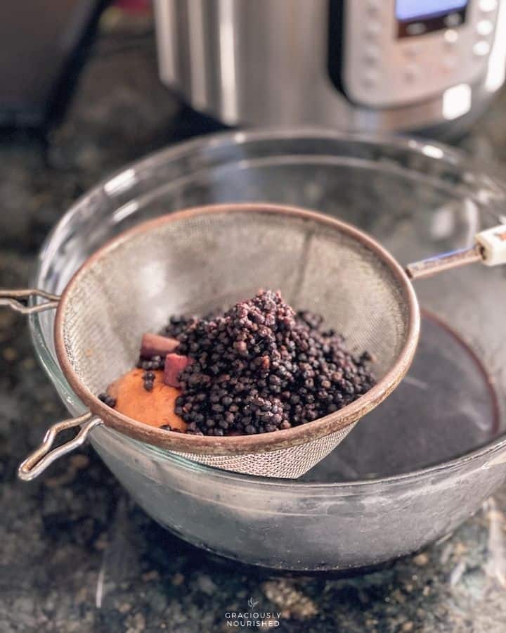 fine mesh strainer with elderberry ingredients over top of a bowl with elderberry syrup. Instant pot in the background. 