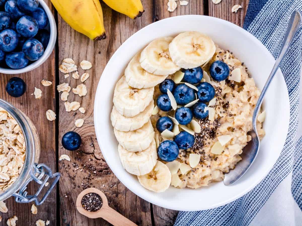 Top down view of oatmeal in bowl topped with blueberries, slivered almonds and banana. Bananas, b blueberries and oats on table. 