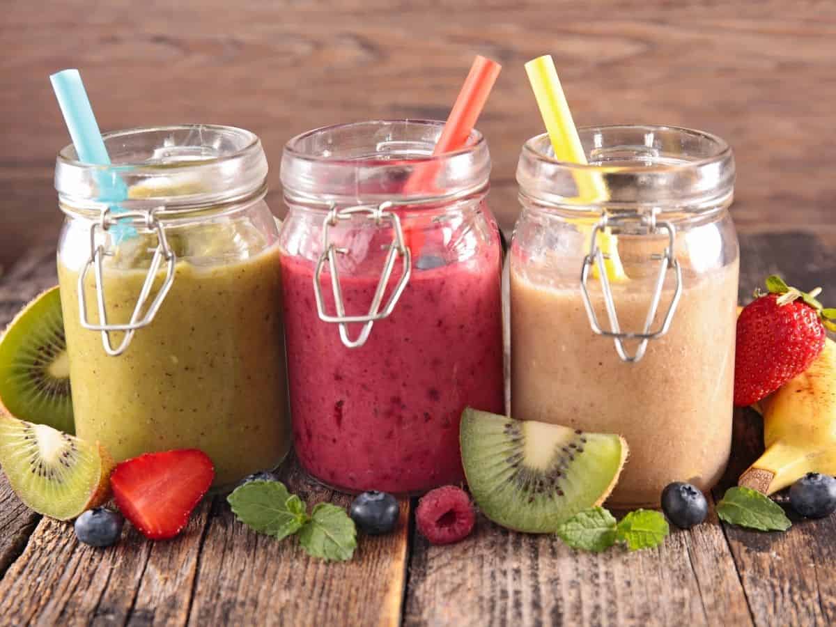 three smoothies in mason jars next to each other. Fruit around them on table.