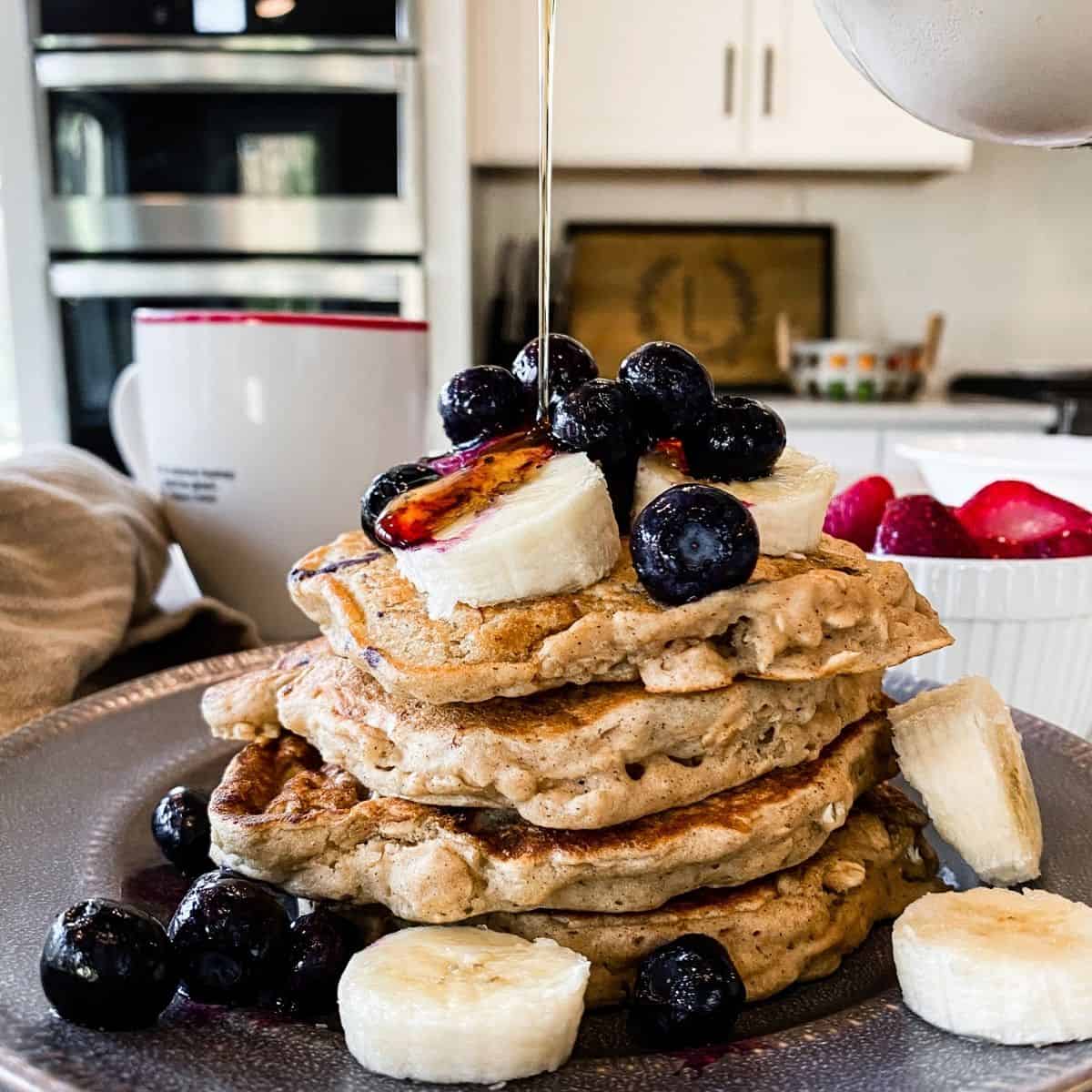Syrup being poured over a stack of vegan oatmeal banana pancakes topped with bananas, blueberries. 
