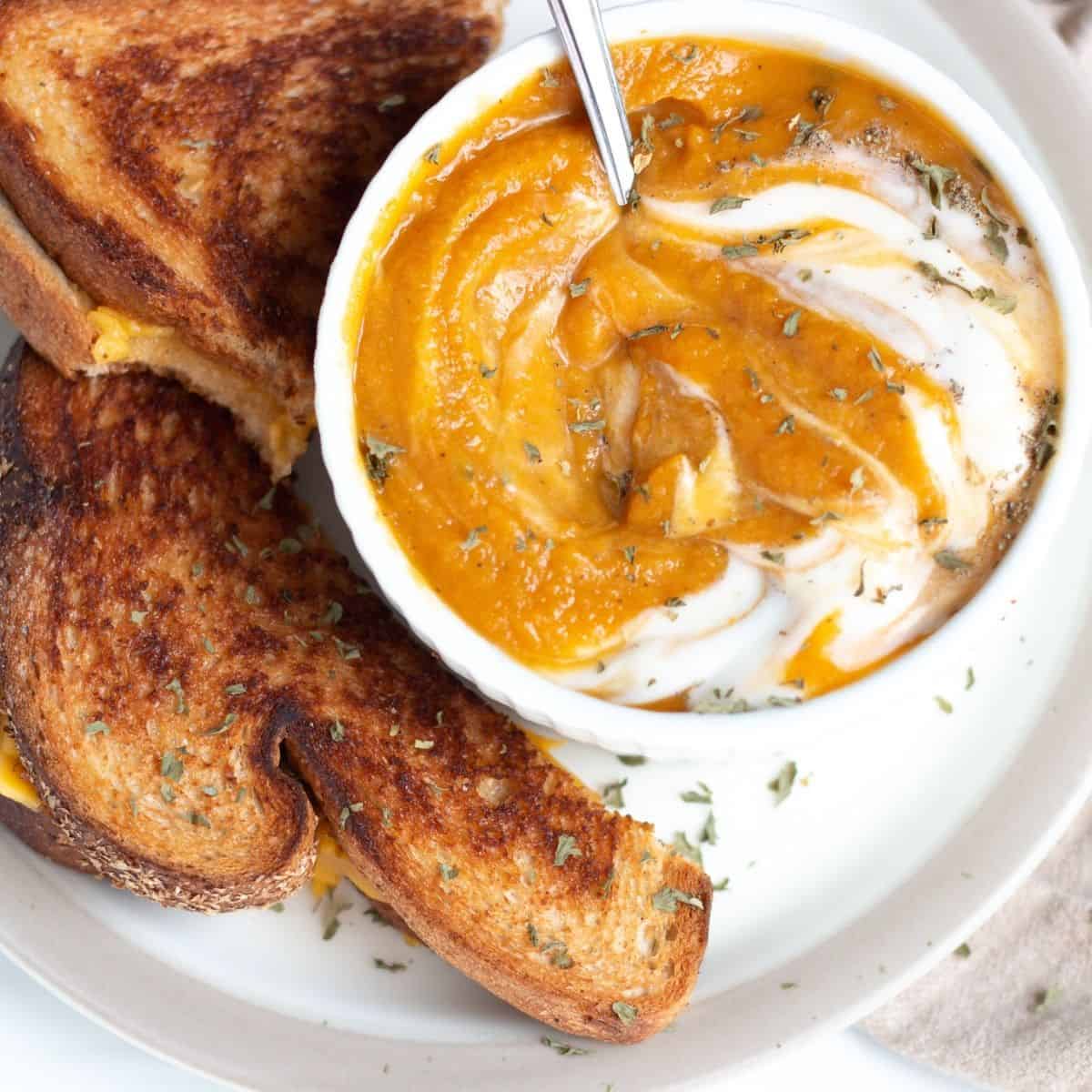 Pumpkin and sweet potato soup with sour cream swirled into it an sage sprinkled on top next to grilled cheese sandwich 