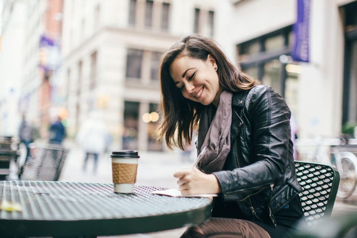 woman sitting outside of a cafe drinking coffee and journaling her intuitive eating hunger scale. 