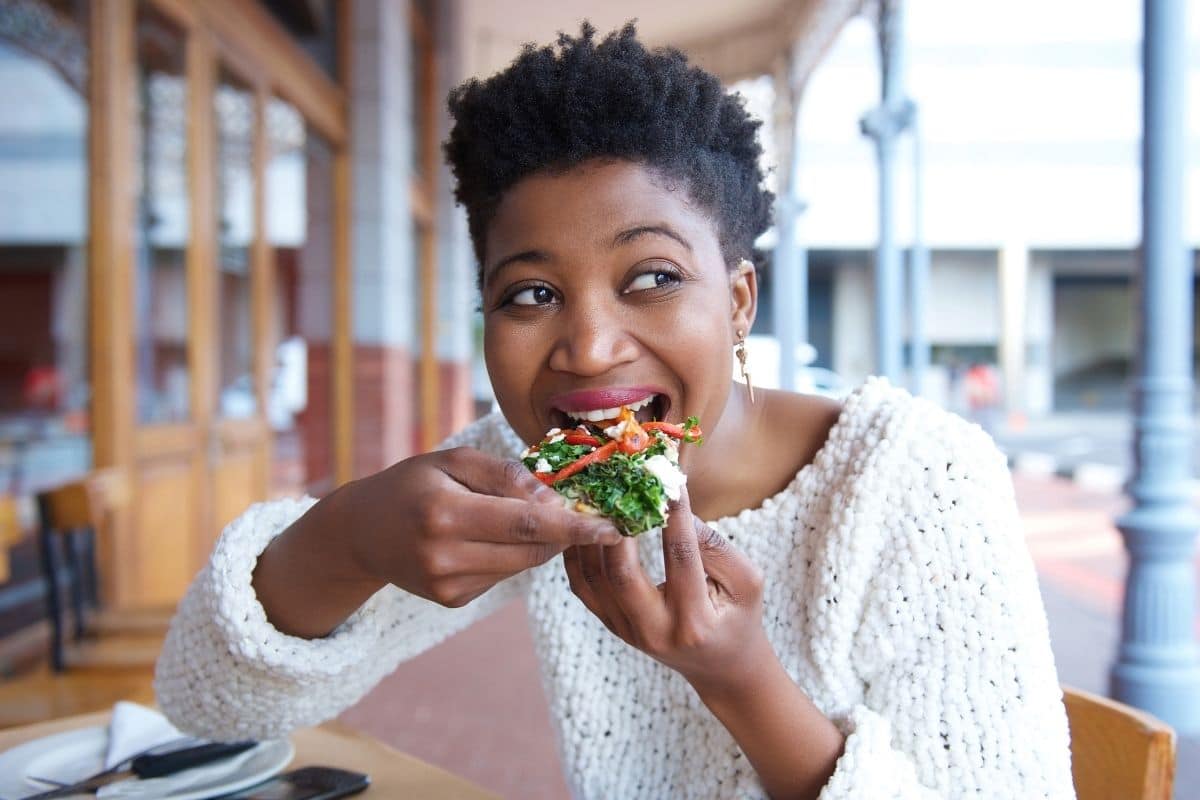 woman sitting outside of a restaurant at a table eating a veggie filled flatbread. 