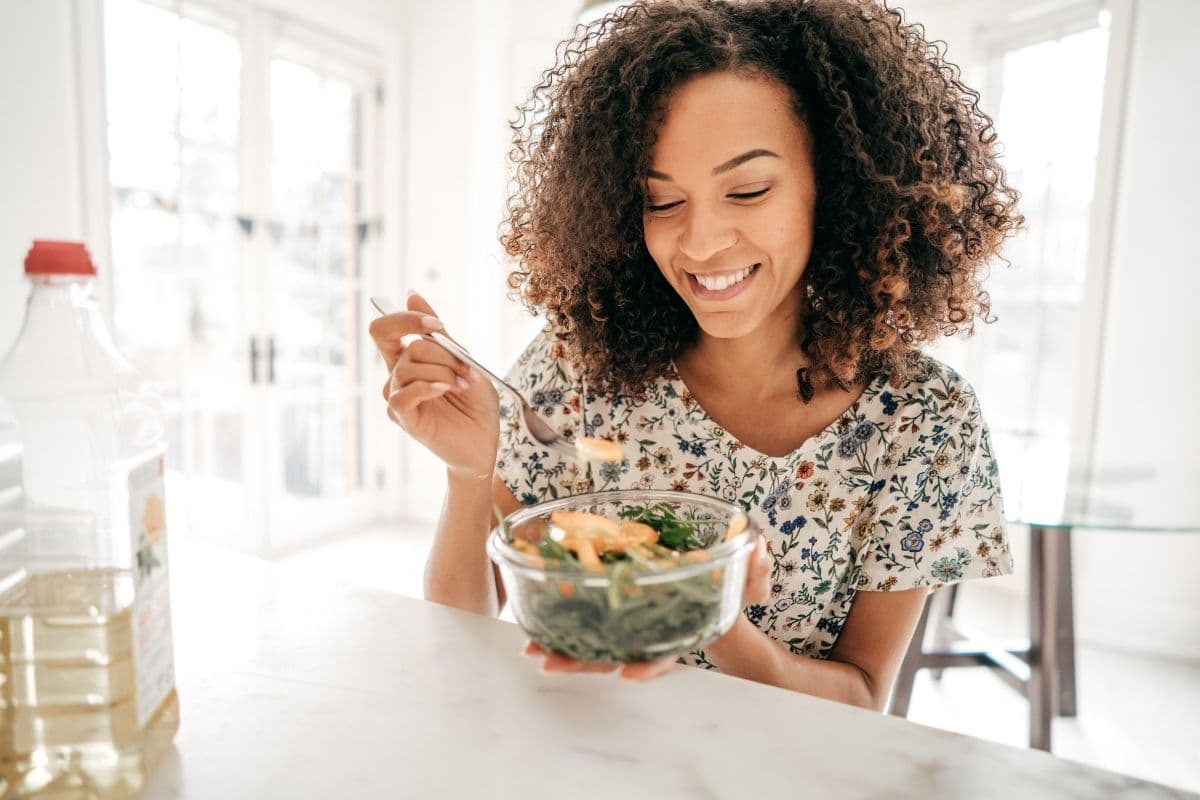 woman smiling wearing a floral shirt and sitting at table eating salad out of a clear bowl. 