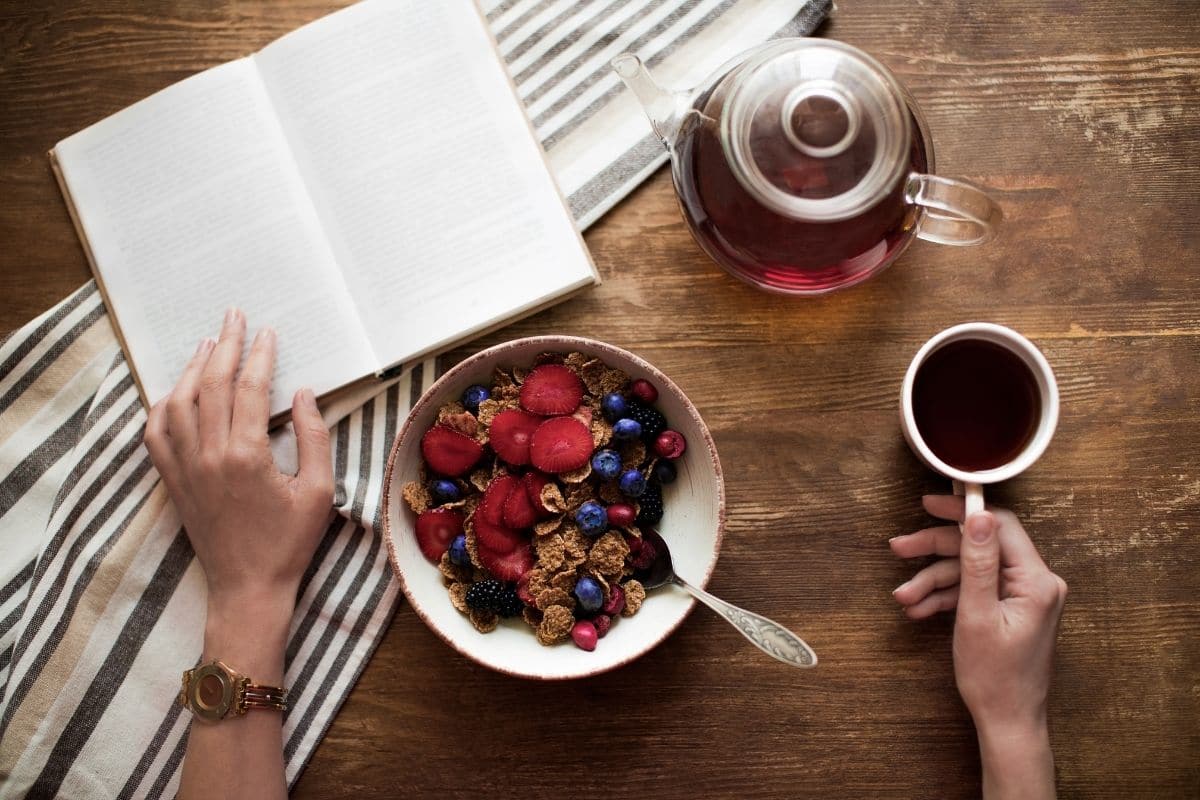 wooden table background with a tea pot, cup of tea, a book, and a bowl of fruit and granola on yogurt. Womans hand holding down a page of the book and the handle for her teacup. 