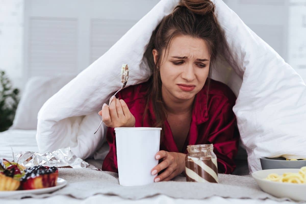 woman sad under white blanket emotionally eating ice cream and other  foods. 