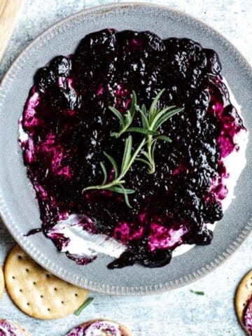 a plate of blueberry goat cheese garnished with rosemary leaves on a concrete table. surrounded by crackers slathered with dip.