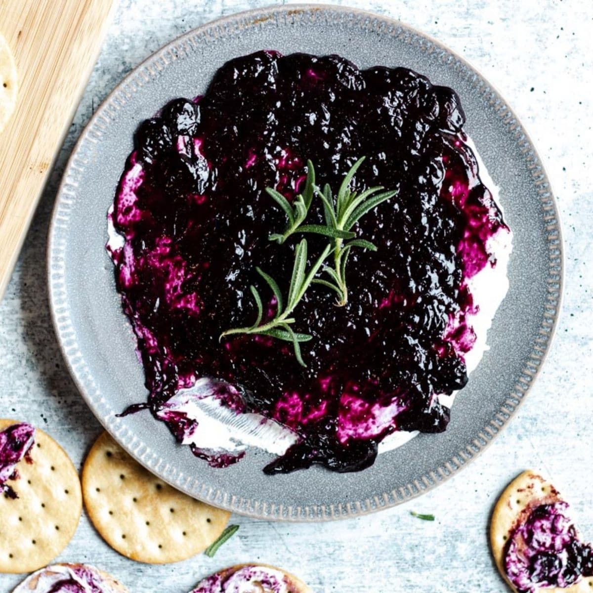 a plate of blueberry goat cheese garnished with rosemary leaves on a concrete table. surrounded by crackers slathered with dip.