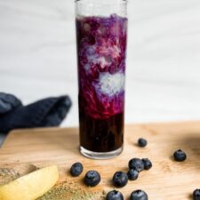 iced blueberry matcha latte in a tall glass on a wooden cutting board. Blueberries and lemon on the cutting board in front of the latte.
