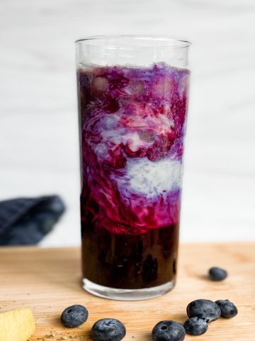 blueberry matcha latte in a tall glass with swirls of white and purple. Sitting on a wooden cutting board with lemon wedges and blueberries.