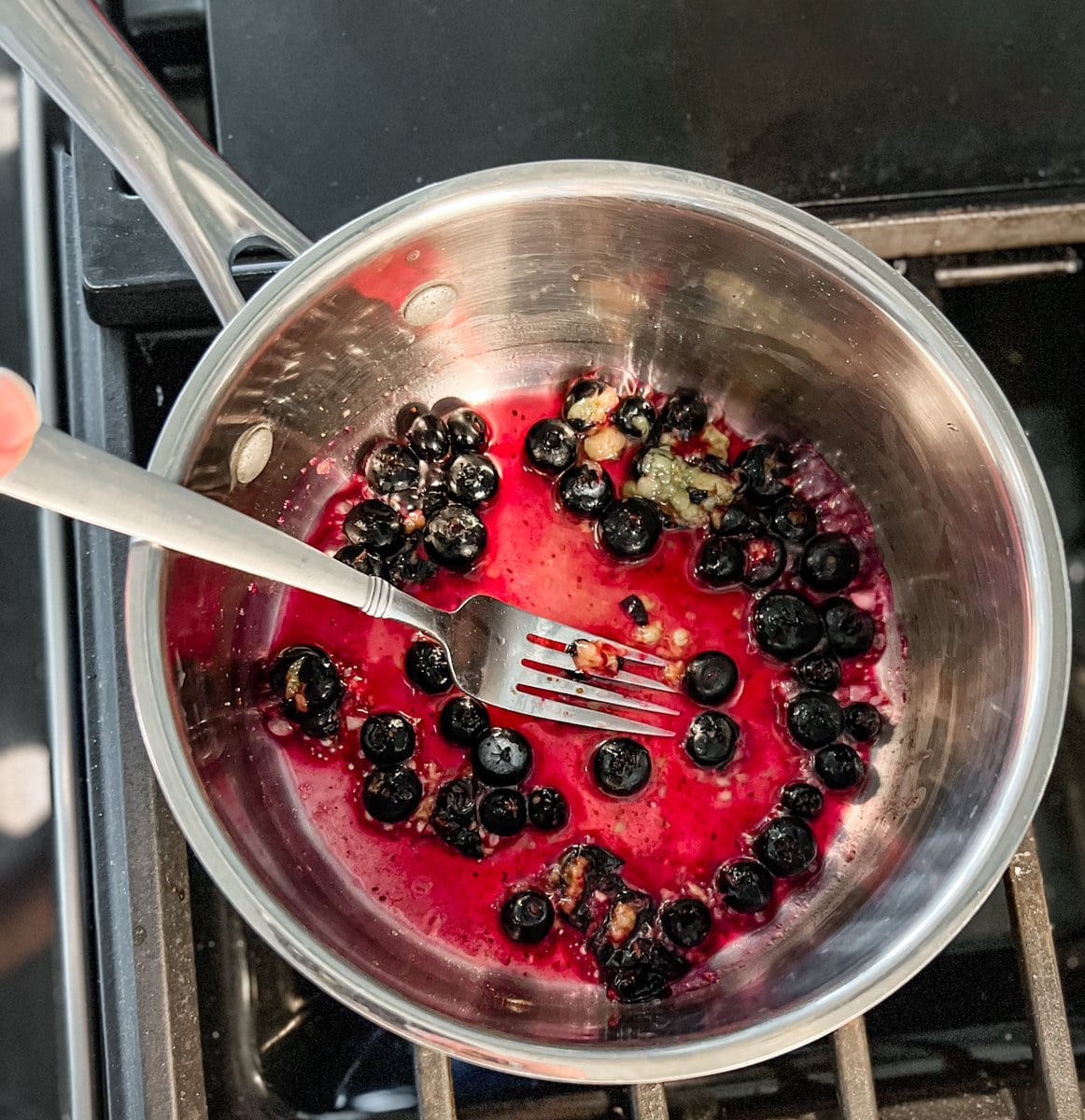 a stainless steel saucepan on a gas stove with blueberries cooking inside. a silver fork gently smashing the blueberries so they release their juices. 
