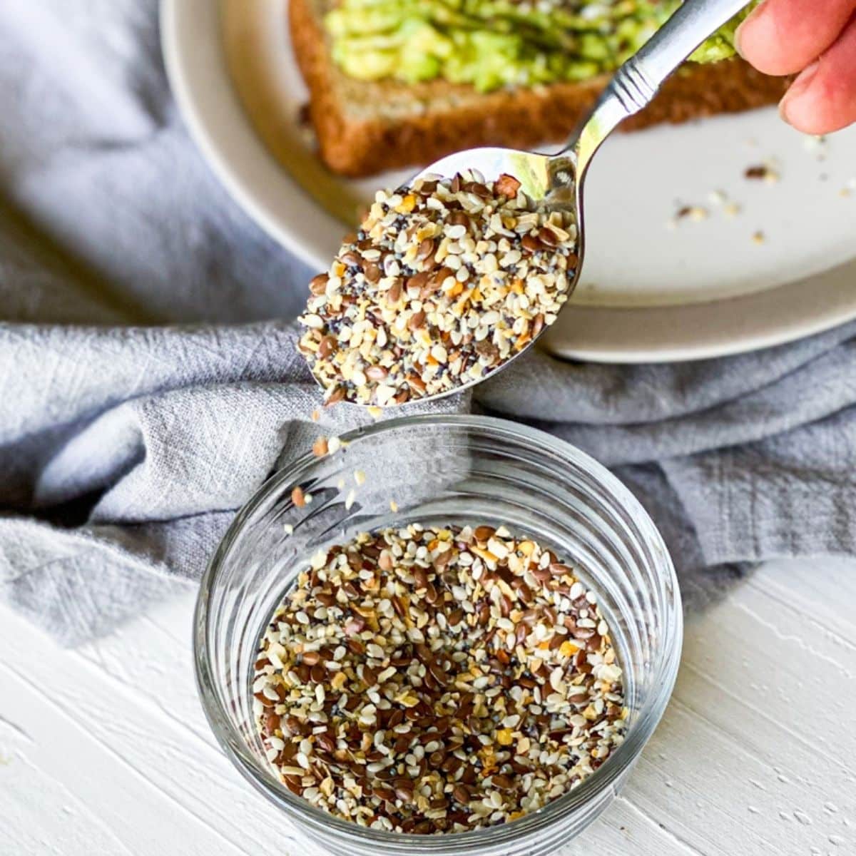 everything bagel seasoning in a small dish with a spoon scooping seasoning and sprinkling it back in. A gray napkin and plate of toast with avocado and seasoning on top in the background. 