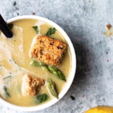 top down view of bowl of soup with croutons asparagus spears and a lemon wedge on the concrete table next to it.