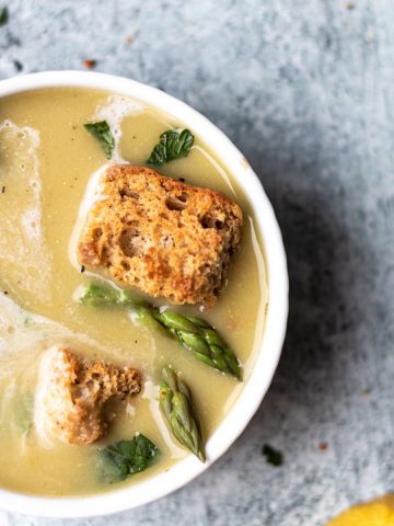 bowl of asparagus soup topped with croutons, coconut milk, and asparagus spears in a white bowl on top of a concrete background.