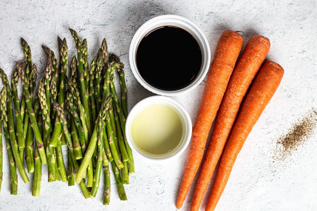 Ingredients for roasted carrots and asparagus- balsamic vinegar, asparagus, carrots, olive oil, and salt and pepper on a white background. 