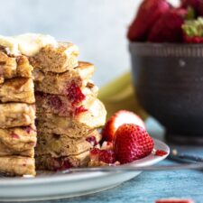 a stack of strawberry banana pancakes on a plate garnished with strawberries. Bowl of berries in the background.