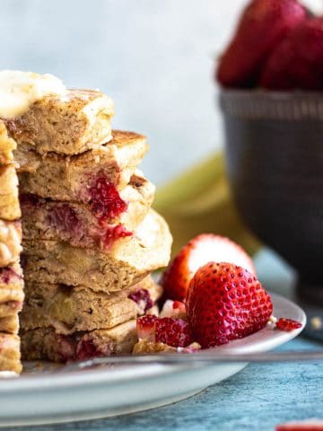 a stack of strawberry banana pancakes on a plate garnished with strawberries. Bowl of berries in the background.