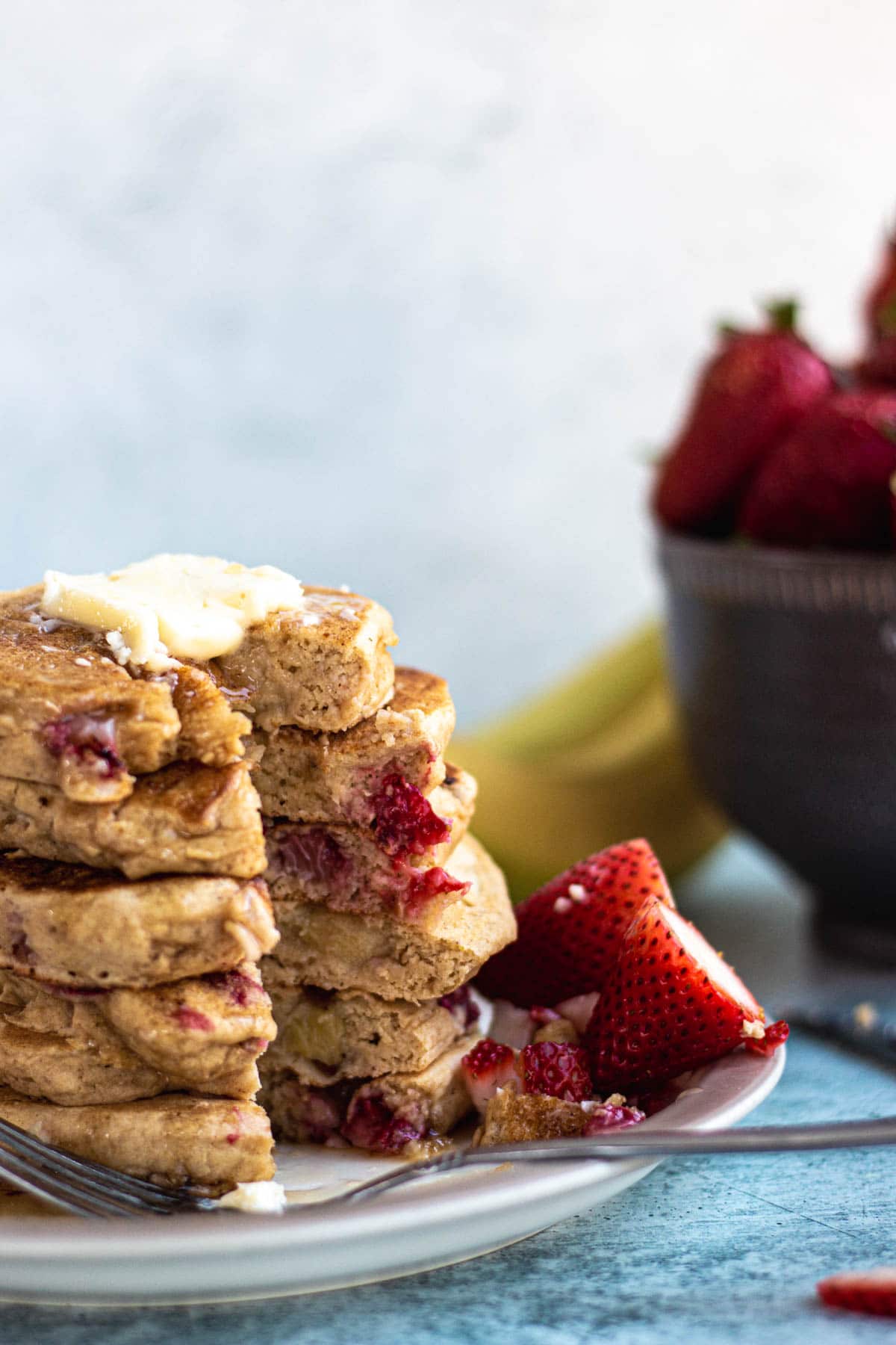 A stack of strawberry banana pancakes on a plate with a pat of butter on top.  Sliced strawberries on the plate. A bowl of strawberries in the background.