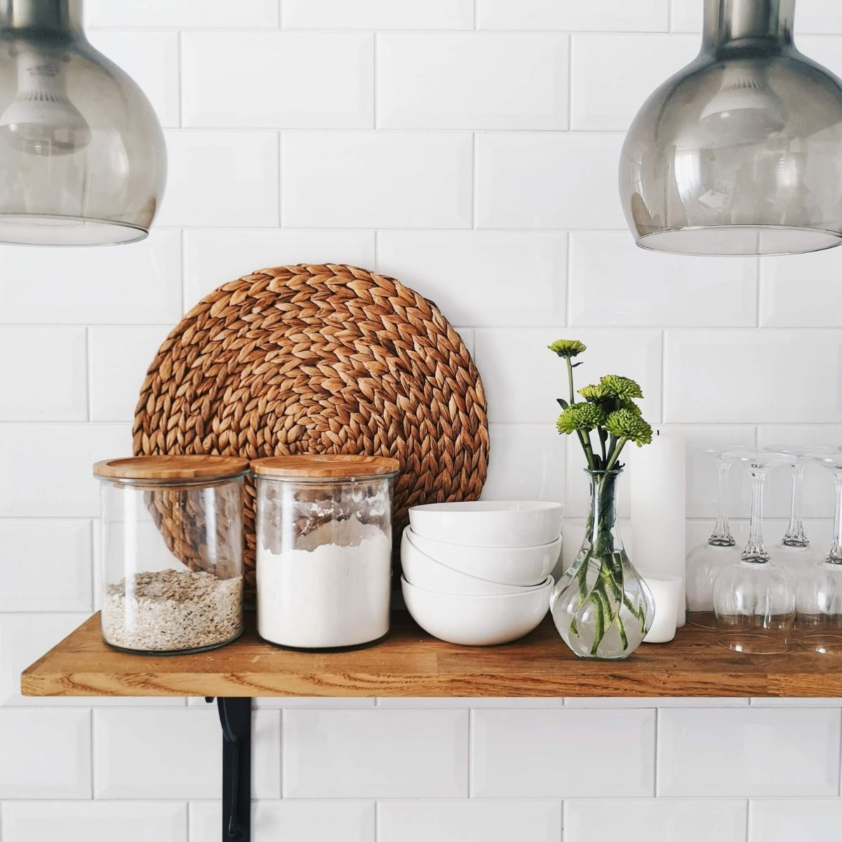 wood shelf on a white wall. Clear jars with wooden tops containing baking ingredients, white bowls, and greens in a vase sitting on shelf.