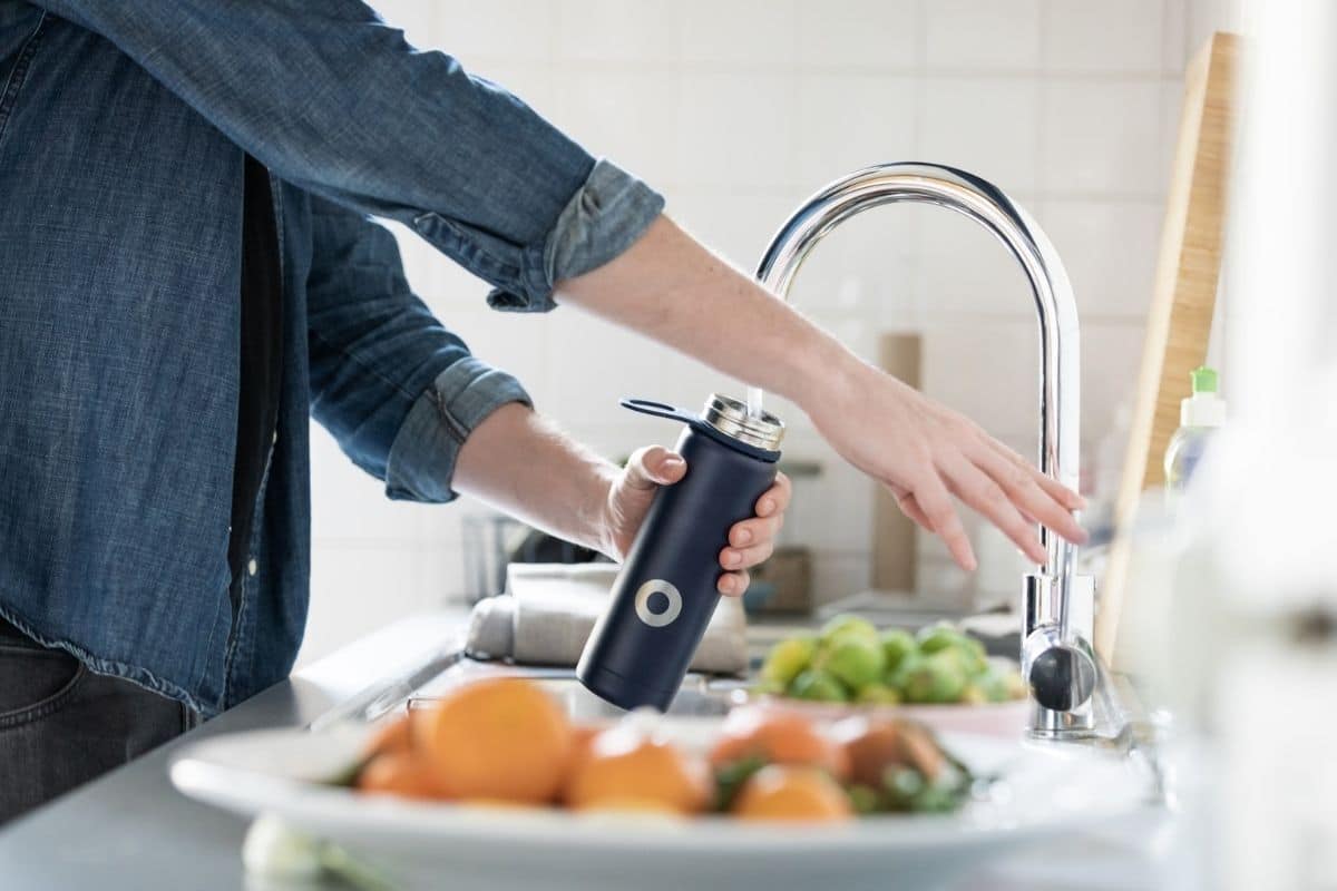 woman filling up a blue steel water bottle in an industrial looking sink with plates of fruit in the foreground and background. 
