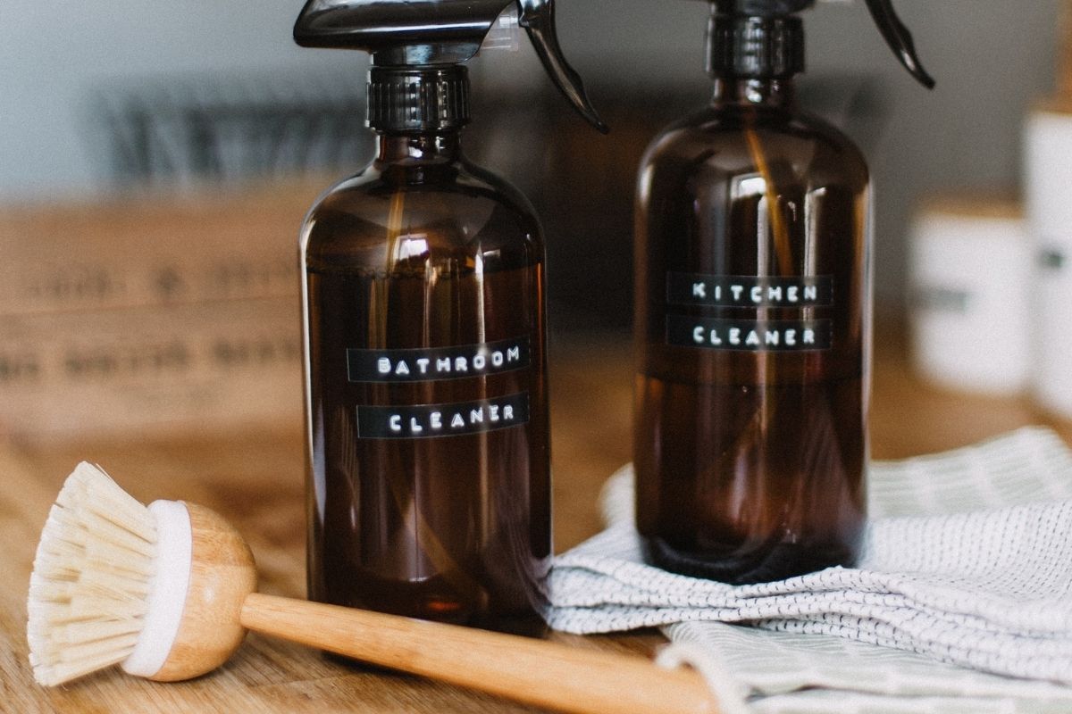 two glass amber bottles with cleaning products sitting on a wooden table alongside  a reusable wooden brush and cleaning rag. 