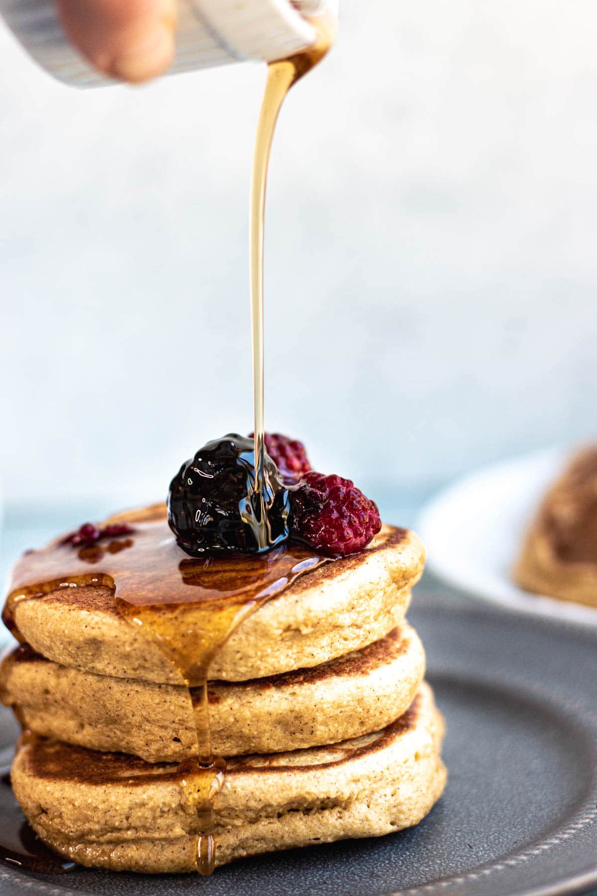 stack of pancakes on a blue plate, topped with blackberries and raspberries. Syrup drizzling on top. 