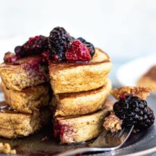 stack of 3 pancakes with berries on top and a small section cut out. all sitting on a blue plate.