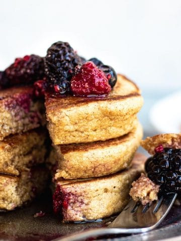 stack of 3 pancakes with berries on top and a small section cut out. all sitting on a blue plate.