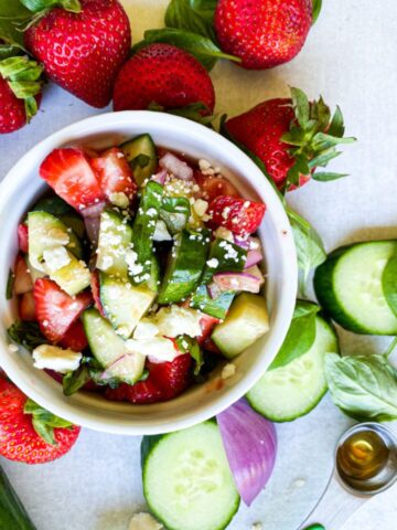 cucumber strawberry salad in a white bowl surrounded by whole strawberries, sliced cucumbers, basil leaves, and red onion slices.
