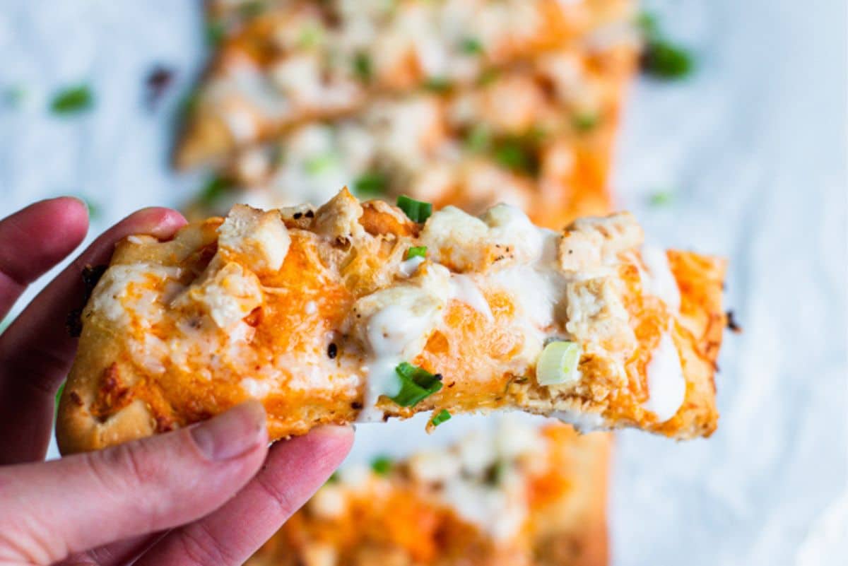 a hand holding a piece of buffalo chicken flatbread close to the camera. 