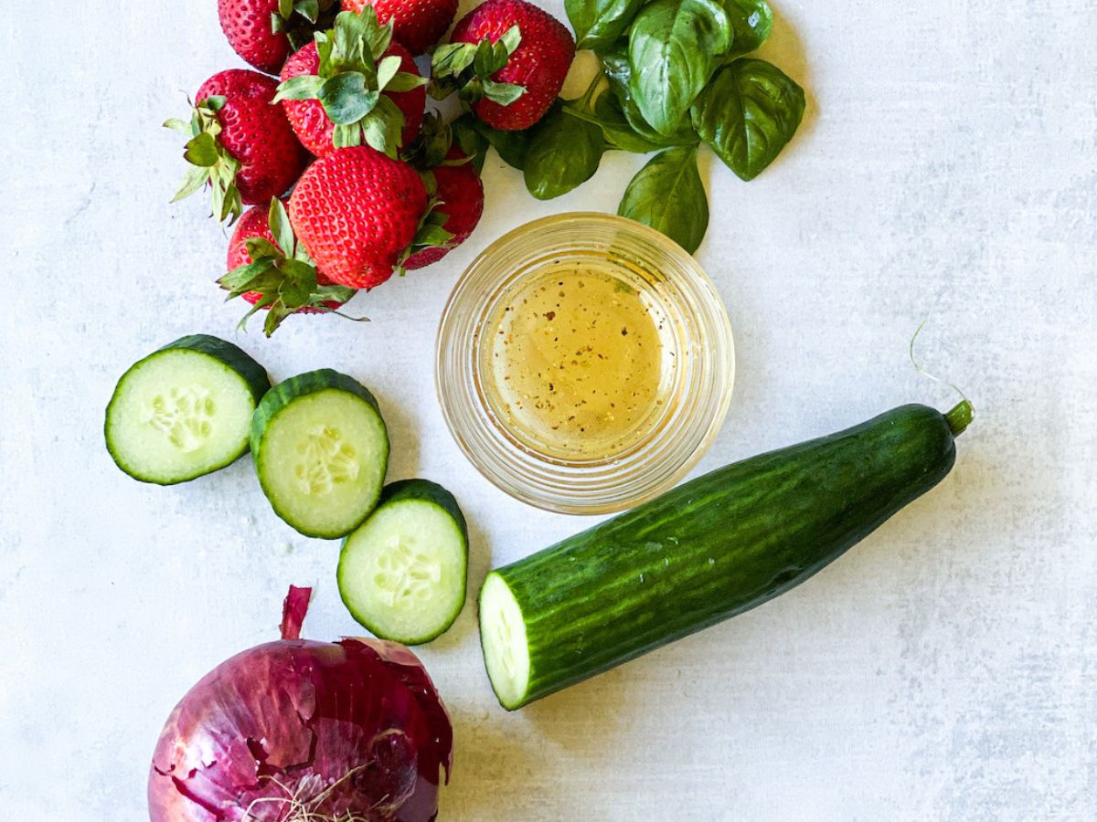 strawberries, cucumber slices, honey vinaigrette, basil, and cucumber on a white table. 