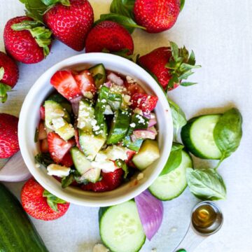 cucumber strawberry salad prepared in a white bowl surrounded by cucumber, strawberries, basil, and red onion
