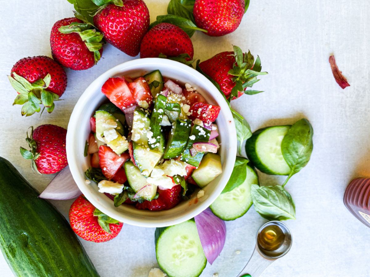 cucumber strawberry salad prepared in a white bowl surrounded by cucumber, strawberries, basil, and red onion
