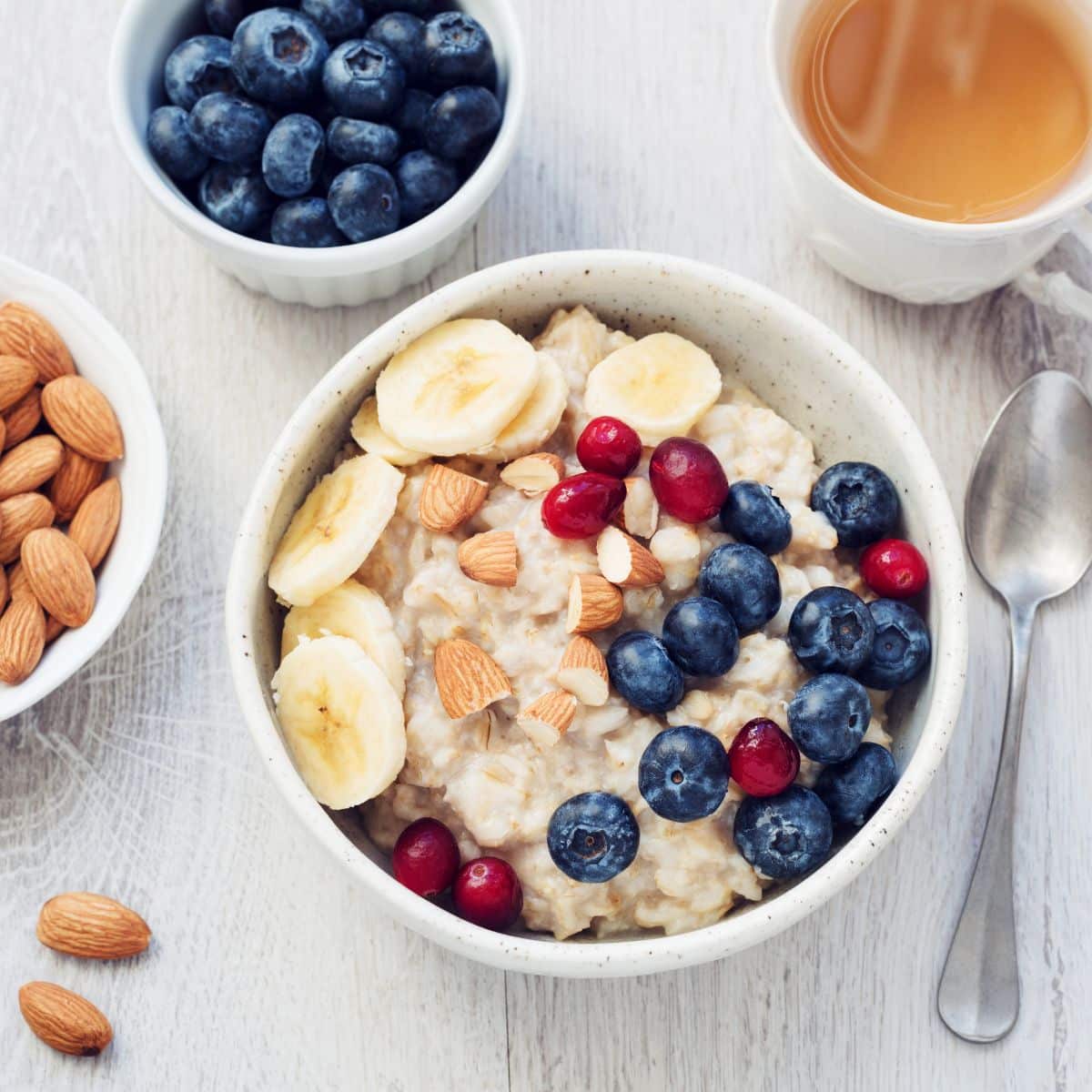 Bowl of oatmeal topped with berries, bananas, and nuts with bowls of berries, almonds, and a cup of tea. 