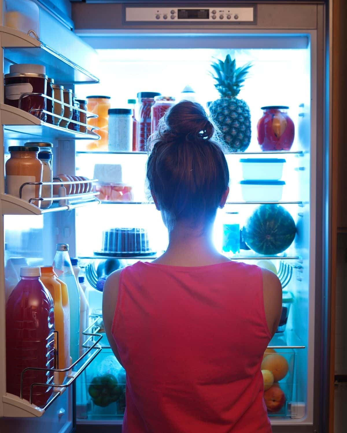 Woman standing in front of the fridge at night, looking inside while deciding on a snack.