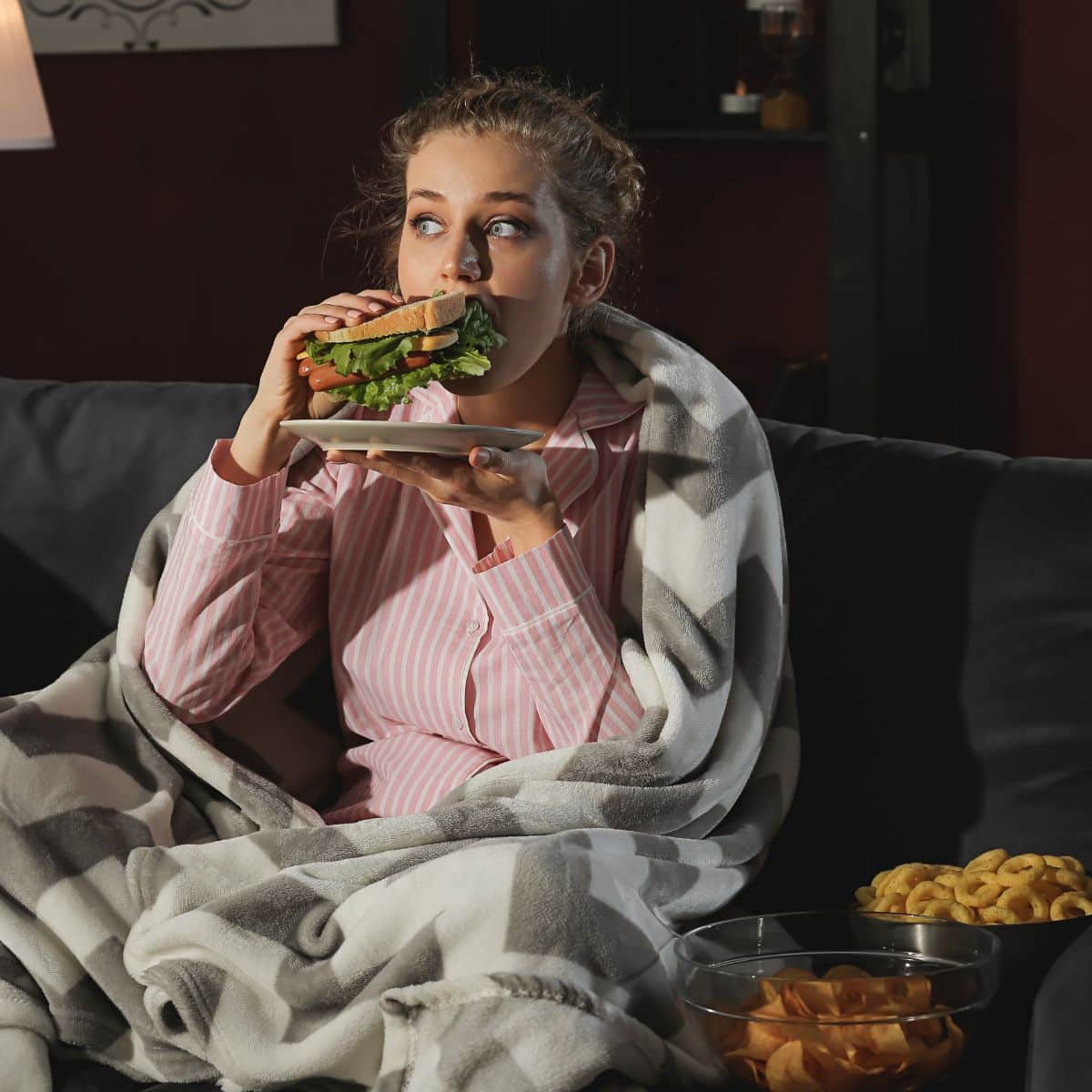 woman sitting on the couch at night with snacks around her, eating a sandwich.