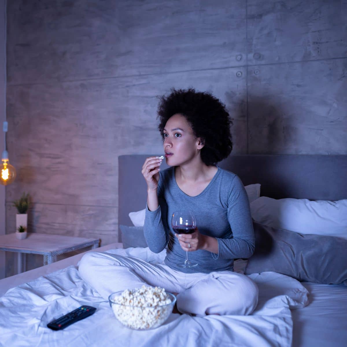 woman sitting in bed watching tv with a bowl of popcorn and a glass of wine. 