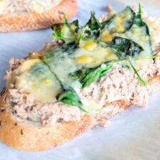 An open face tuna melt topped with melted cheddar cheese, served on whole-grain bread with baby spinach on a white plate.