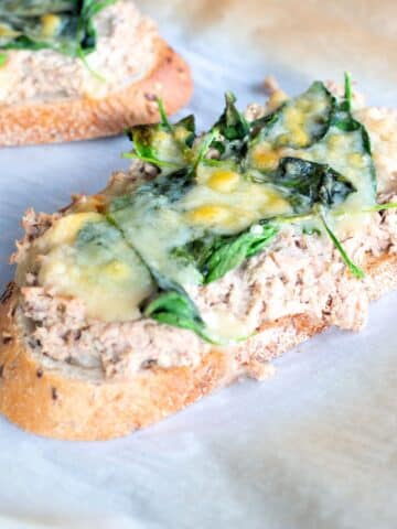 An open face tuna melt topped with melted cheddar cheese, served on whole-grain bread with baby spinach on a white plate.
