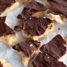 Frozen chocolate peanut butter banana bark with layers of mashed banana, peanut butter, and melted chocolate, topped with optional toppings like sea salt and crushed peanuts, served on parchment paper.