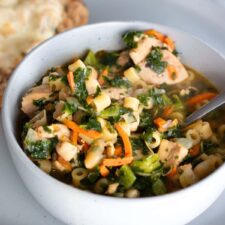 A bowl of white bean and chicken soup with kale, carrots, and ditalini pasta, garnished with fresh herbs, served with a slice of crusty bread on the side.
