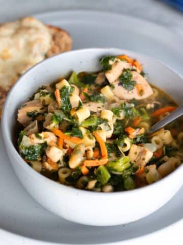 A bowl of white bean and chicken soup with kale, carrots, and ditalini pasta, garnished with fresh herbs, served with a slice of crusty bread on the side.