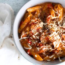 A white bowl filled with Caprese penne pasta, made with penne, sliced pre-cooked chicken sausages, petite diced tomatoes, mozzarella cheese, and a drizzle of balsamic glaze.