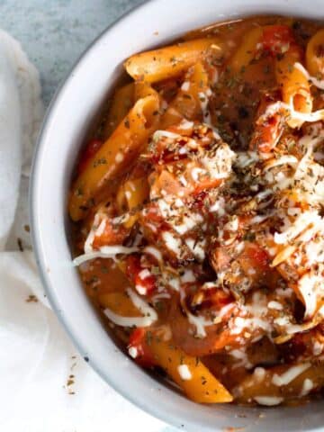 A white bowl filled with Caprese penne pasta, made with penne, sliced pre-cooked chicken sausages, petite diced tomatoes, mozzarella cheese, and a drizzle of balsamic glaze.