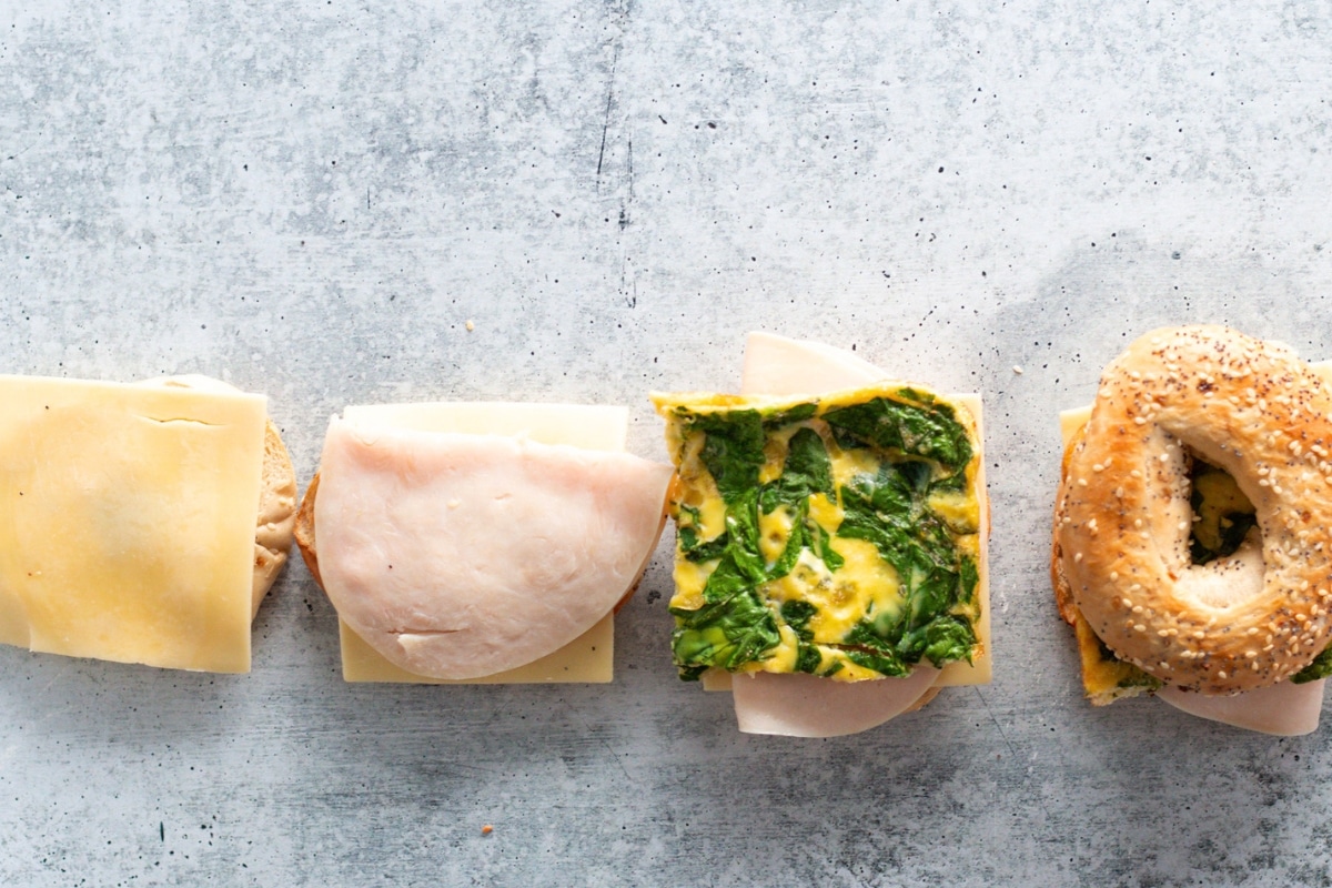 Prepping a healthy breakfast bagel sandwich with eggs, spinach, turkey, and cheese on a sliced everything bagel.