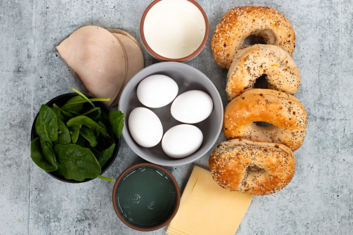 Ingredients for a healthy breakfast bagel laid out on the table, including bagels, eggs, spinach, turkey, cheese, and seasonings.