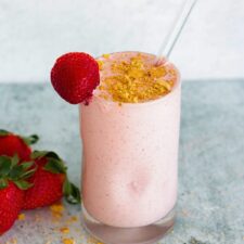 Strawberry cheesecake protein shake in a glass, topped with crushed graham crackers, with fresh strawberries in the background.