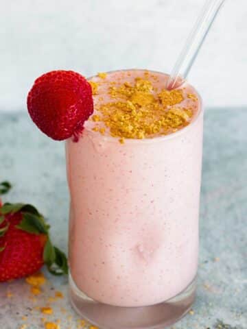 Strawberry cheesecake protein shake in a glass, topped with crushed graham crackers, with fresh strawberries in the background.