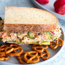 A salmon salad sandwich made with Greek yogurt and healthy ingredients, served on a white plate with a side of pretzels.